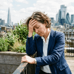 Gestresste Frau auf Dachterrasse in London, hält sich den Kopf, zeigt Anzeichen von Belastung und Stress, mit moderner Skyline im Hintergrund.